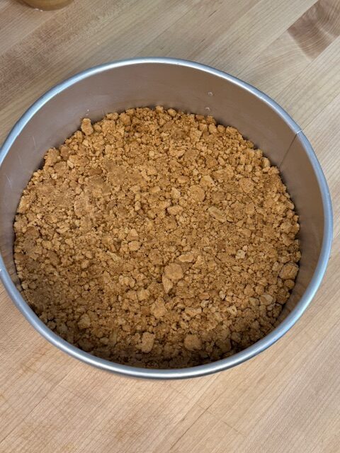Crushed nutter butters mixed with melted butter pressed into the bottom of a metal cake pan on a wooden cutting board.