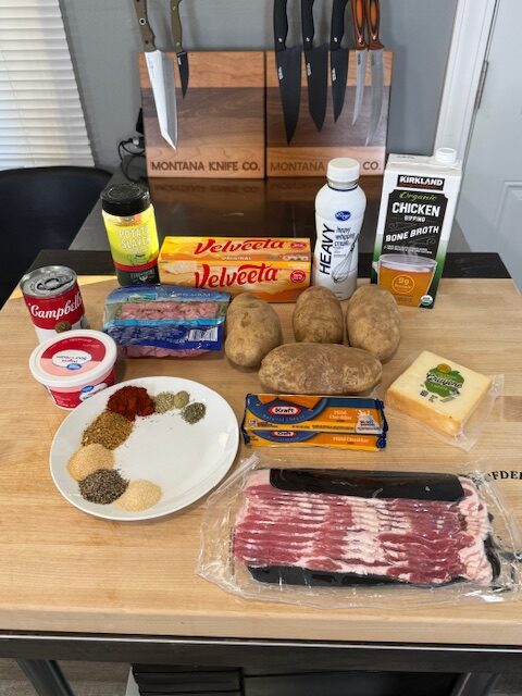 Ingredients displayed on wood cutting board for slow cooker cheesy potato soup. Ingredients include potatoes, chicken broth, ham, heavy cream, sour cream, cream of chicken, gruyere cheese, Velveeta cheese, bacon, potato slayer seasoning, garlic powder, smoked paprika, onion, rosemary, and dried sage.
