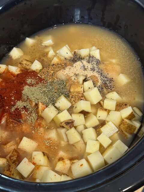 Adding the potatoes, chicken broth, ham, and spices to the black slow cooker insert.
