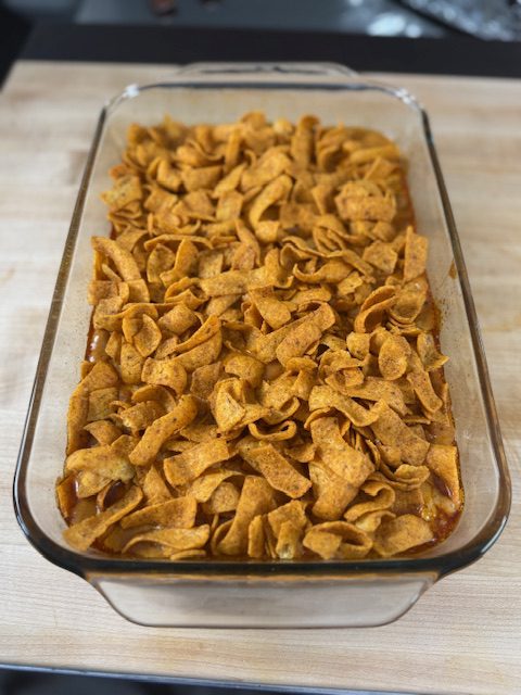 This is the enchilada casserole in a glass baking dish on a wooden counter. There are Fritos on top of the casserole.