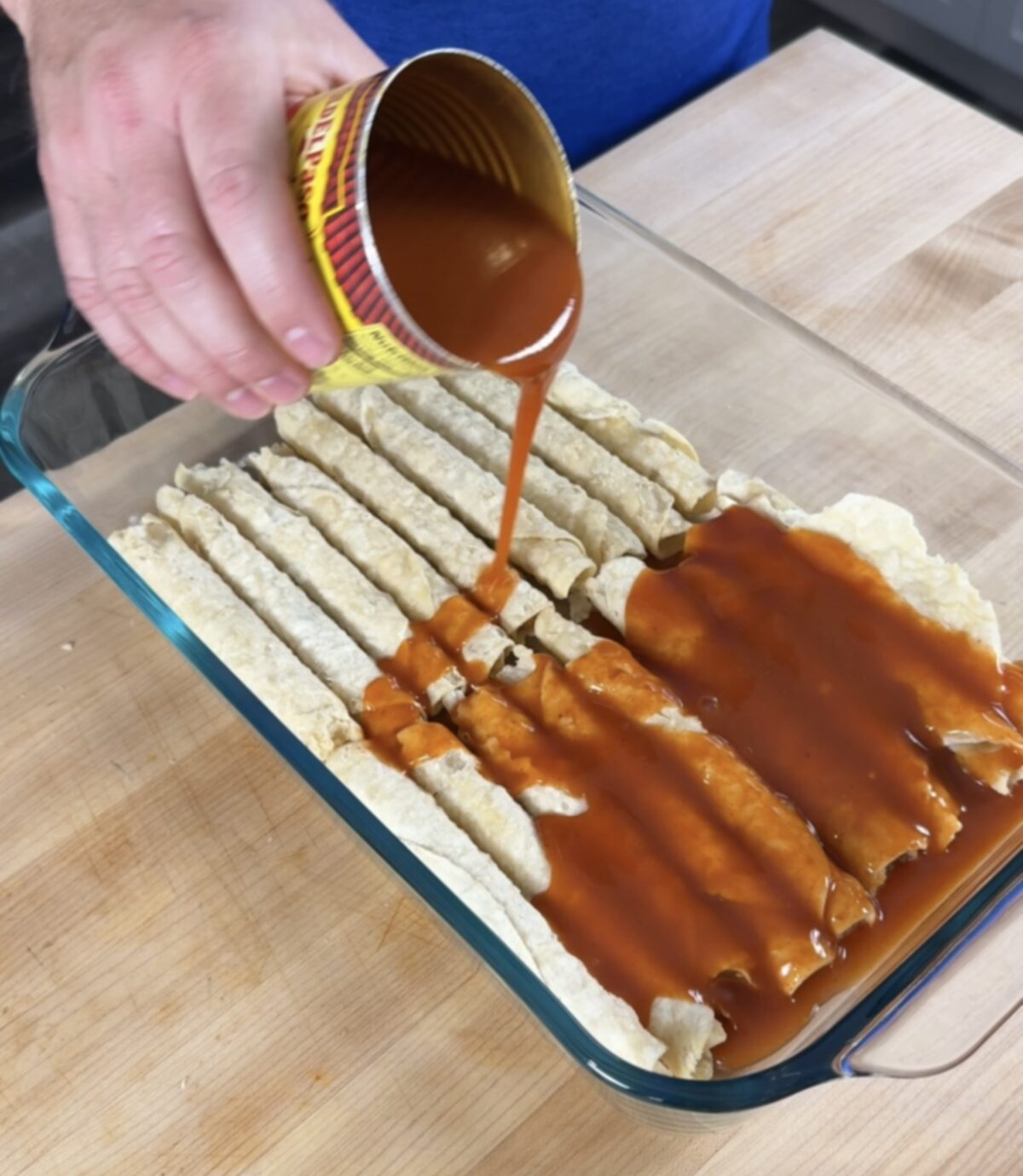 In a glass baking dish is red enchilada sauce and frozen chicken taquitos. A hand is pouring more red enchilada sauce on top of the layered taquitos. 