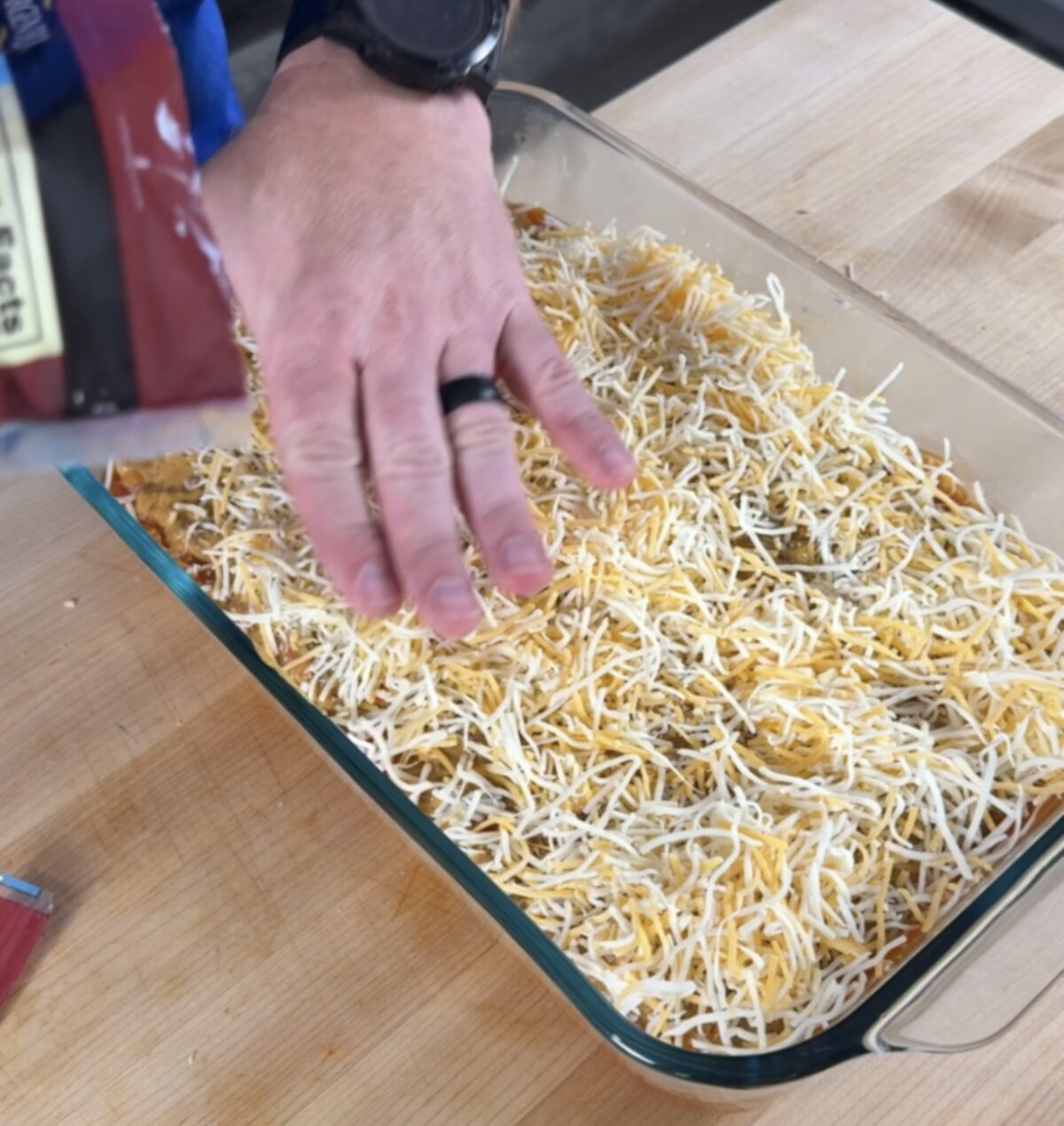 The glass baking dish is topped with ground beef mixture and lots of shredded cheese. Hand is pressing shredded cheese into the top of the casserole.