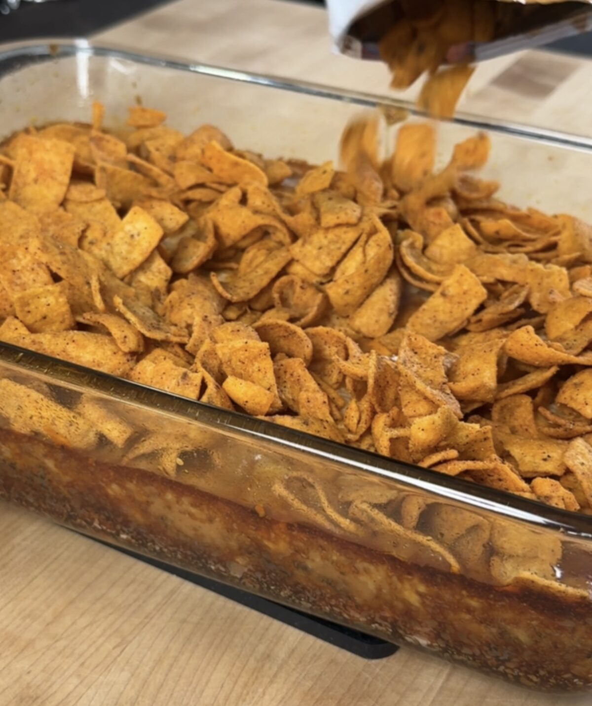 The lazy enchilada casserole is baked and removed from the oven. A hand tops the casserole with Frito chips before serving. 