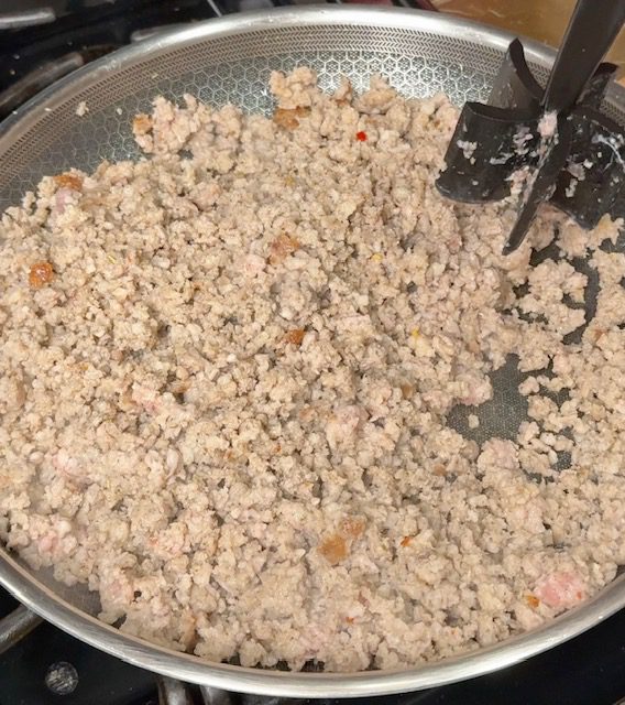 Sausage is being browned in a pan and crumbled with a meat masher.