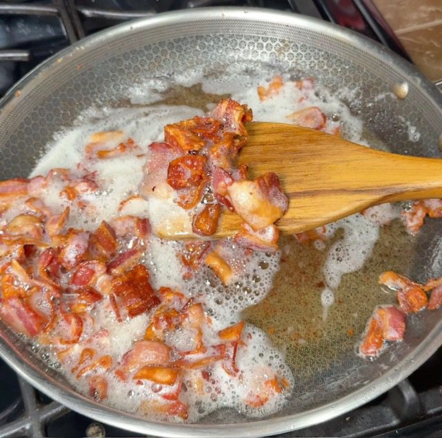 Bacon is being cooked in a pan. A spatula is presenting cooked bacon.