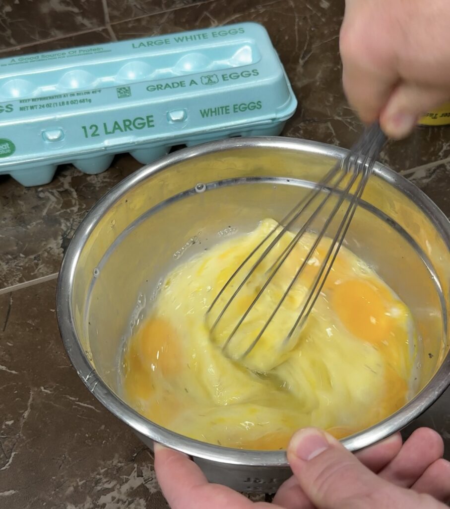 Stainless steel bowl on counter with eggs being beaten with a whisk. Egg carton in background.