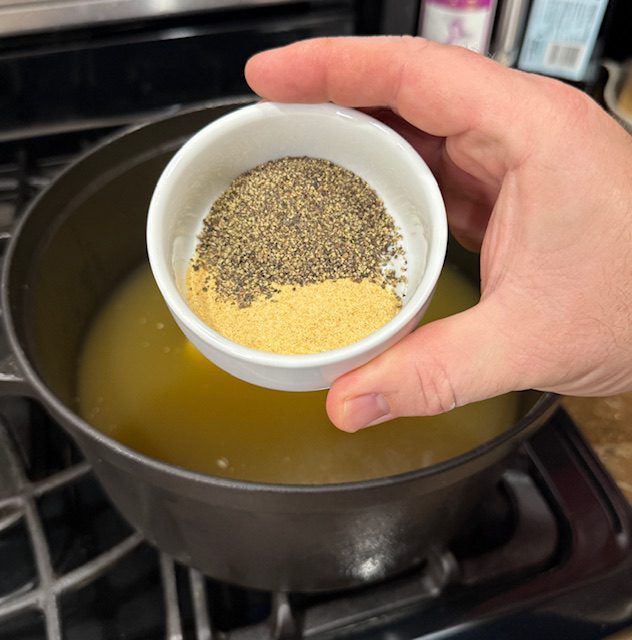 Hand hold a white ramekin of garlic powder, pepper and salt, being added to large pot with broth inside. 