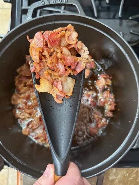 Bacon being cooked until golden brown in a large black pot. Some of the cooked bacon is being show in a spoon.