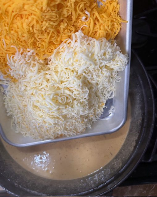 Shredded cheddar cheese and Monterey Jack cheese are on a baking sheet ready to be added to the soup in the pot. 