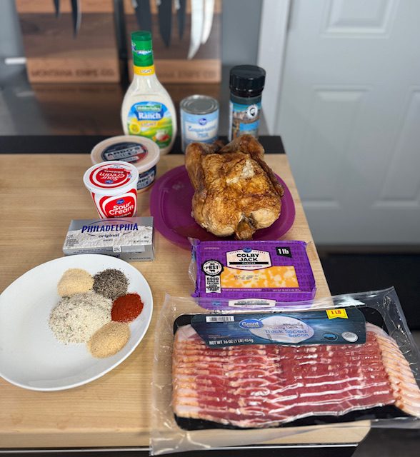 Ingredients displayed on the wood kitchen counter for slow cooker crack chicken dip. Ingredients include, cream cheese, colby jack cheese, bacon, sour cream, cheese dip, ranch seasoning, garlic powder, onion powder, smoked paprika, black pepper, onion powder, and evaporated milk. A rotisserie chicken is also on a plate. 