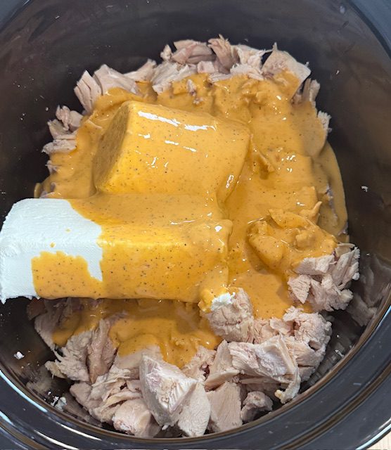 Chicken, cream cheese, and cheese dip are added to the black slow cooker bowl. A white spatula is also in the bowl.