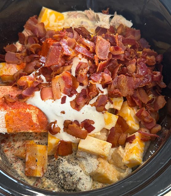 In the black slow bowl is chicken, cream cheese, cheese dip, cube colby jack cheese, and cooked bacon. A white spatula is also in the bowl. 