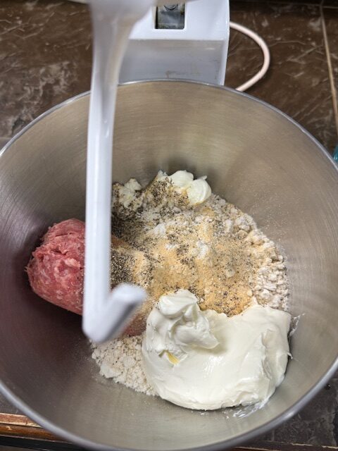 Sausage ball ingredients are being mixed in a stand mixer with a dough hook. The metal mixer bowl has sausage, cream cheese, old bay biscuit mix, garlic powder, salt and pepper.