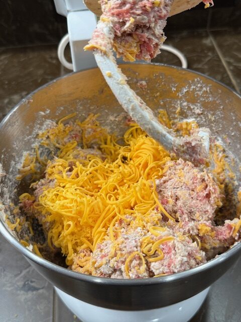 A stand mixer with a metal bowl and a dough hook has the sausage ball mixture and shredded cheddar cheese is added on top.