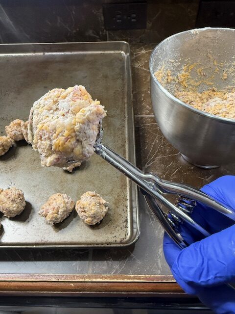 The old bay sausage balls are being scooped onto a baking sheet using a cookie scoop. A hand with a blue glove is holding the cookie scoop and there is the stand mixer bowl with the sausage ball mixture on the side.