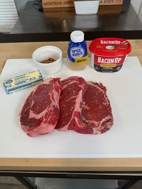 Ingredients for garlic butter steak bites on a plastic cutting board. The ingredient include bacon grease, minced garlic, red pepper flakes, butter, and ribeye steaks.
