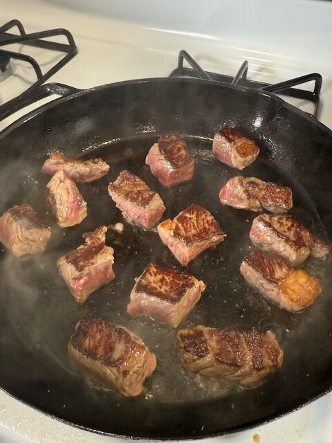 The steak bites have been flipped in the cast iron skillet on the stovetop and are searing on the second side.