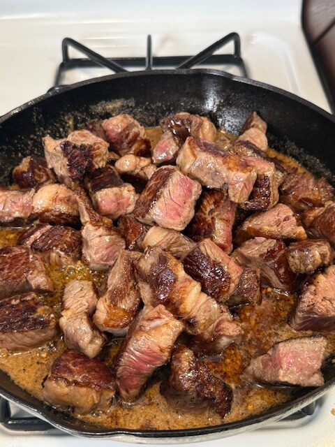 The seared steak bites are added to the garlic butter mixture in the cast iron skillet on the stovetop.