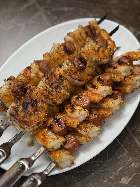Finished shrimp and sausage skewers are grilled and laying on a white plate on a gray counter.