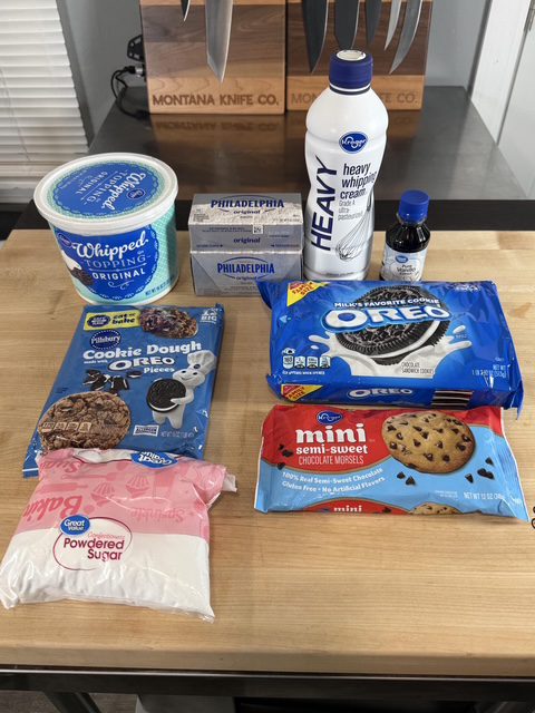 Ingredients for the no-bake Oreo cheesecake are on display on the wooden counter. The products displayed are Oreo cookies, heavy whipping cream, Cool Whip, powdered sugar, chocolate chips, and Oreo cookie dough.