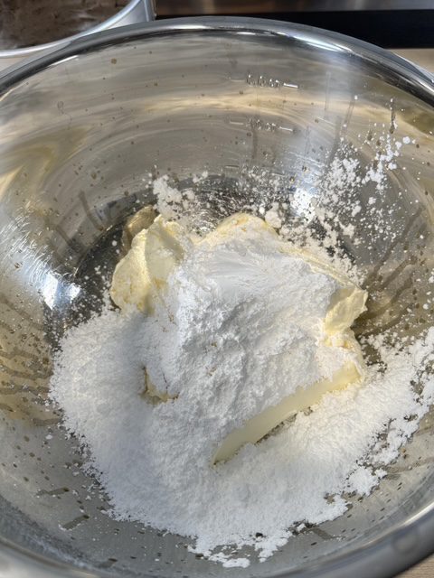 A metal bowl has cream cheese that is being mixed with powdered sugar and vanilla. 