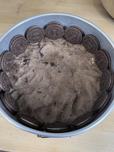 A springform pan as Oreo cookie dough in the bottom of the pan and the sides are lined with Oreo cookies. The pan is on a wooden counter.