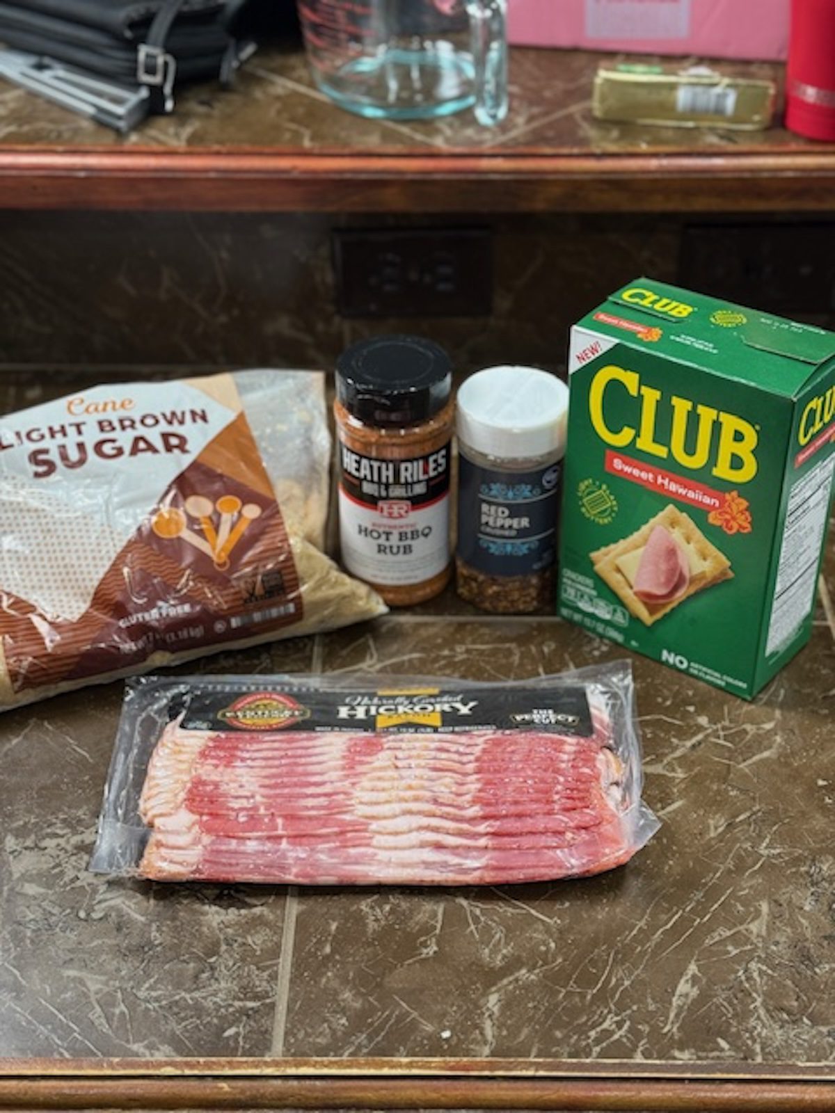 Ingredients for bacon crackers on a counter. The items from left to right include light brown sugar, spicy barbecue rub, bacon, red pepper flakes, and club crackers.