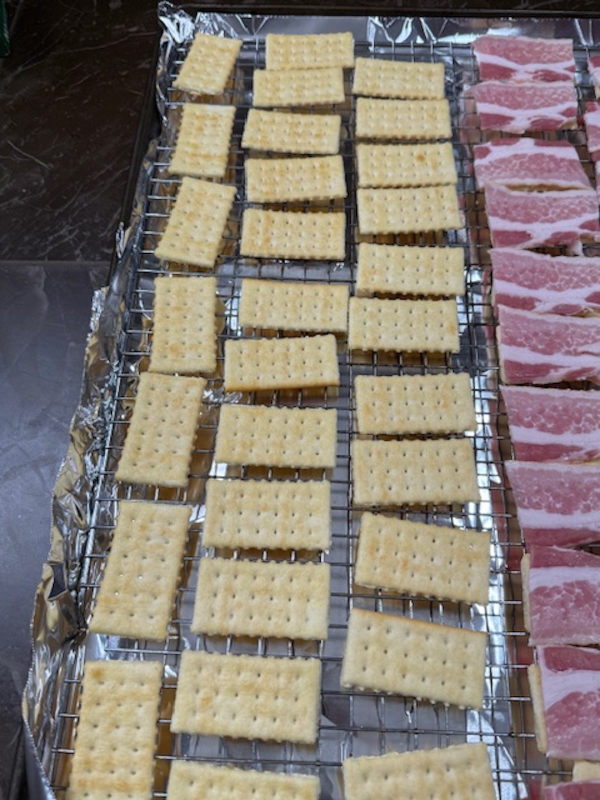 Club crackers and strips of bacon are laid out on a wire rack over a baking sheet lined with foil.