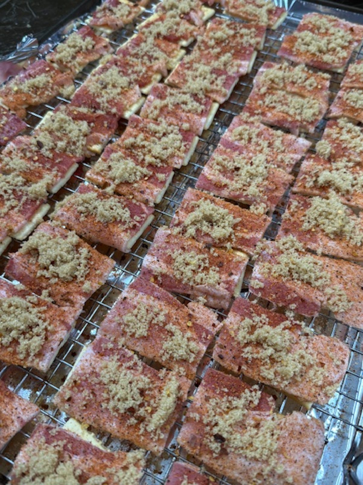 Brown sugar and red chili flakes are sprinkled on top of bacon crackers on a wire rack.