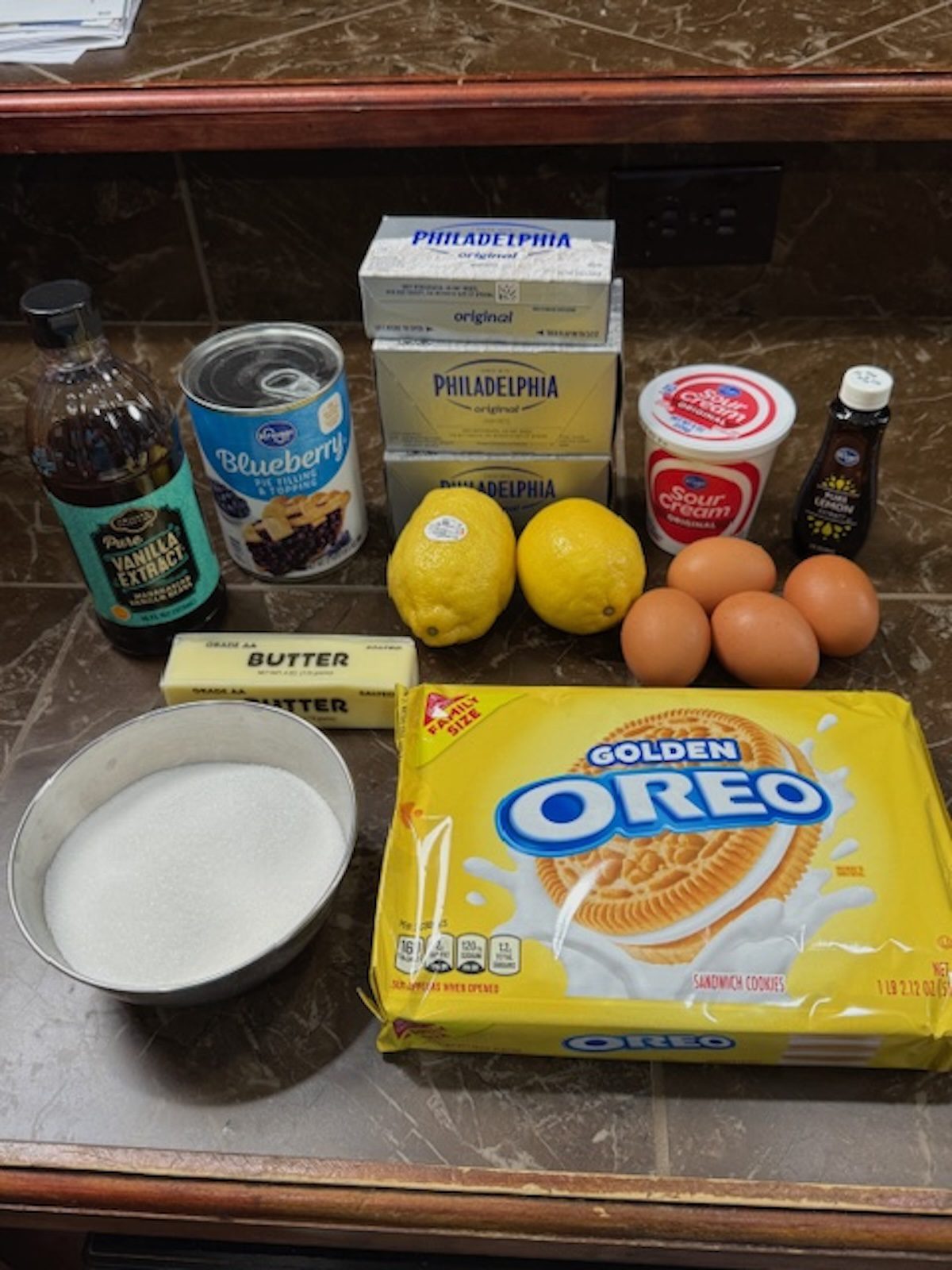 Ingredients for lemon blueberry cheesecake on the counter. Ingredients from left to right include vanilla extract, blueberry pie filling, butter, sugar, cream cheese, lemons, sour cream, eggs, lemon extract, and vanilla Oreos.