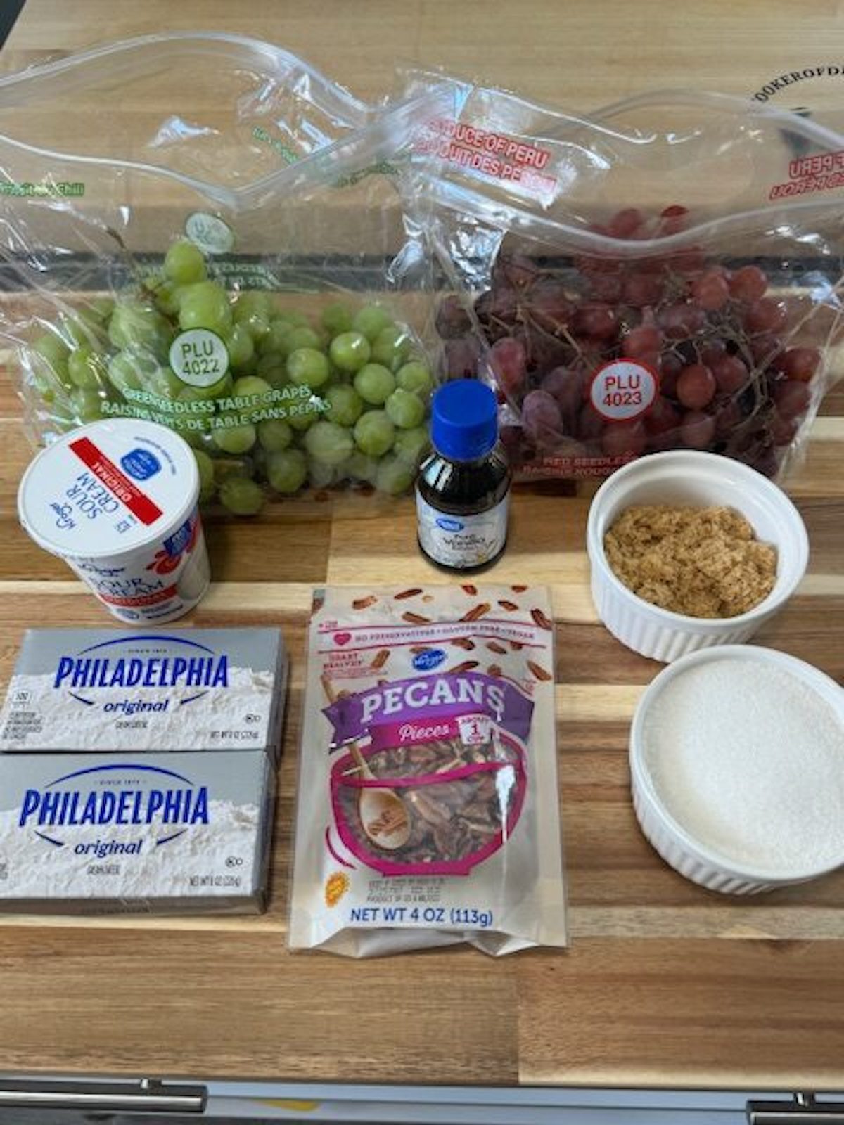 Ingredients for grape salad on a wooden counter. The ingredients include green grapes, red grapes, sour cream, vanilla extract, cream cheese, pecans, light brown sugar, and regular sugar.