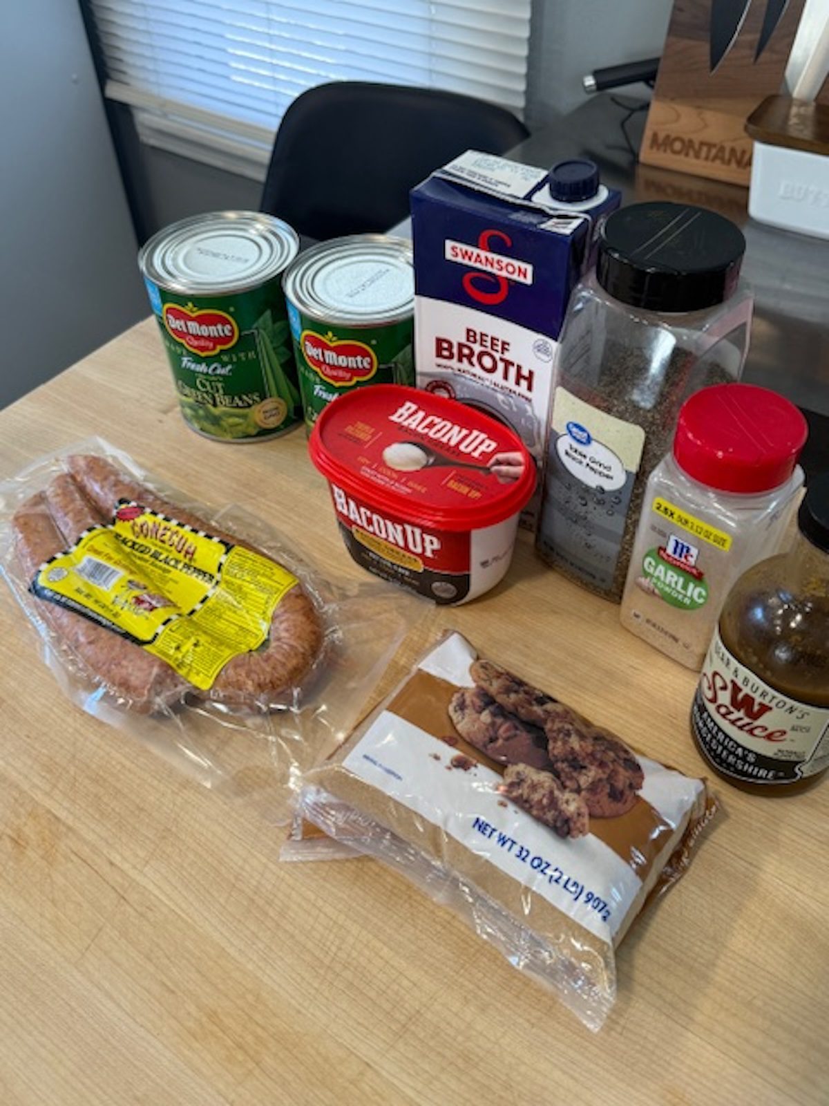 Ingredients for baked green beans on a wood counter. Ingredients include canned green beans, beef broth, pepper, garlic powder, bacon grease, Worcestershire sauce, smoked sausage, and brown sugar.
