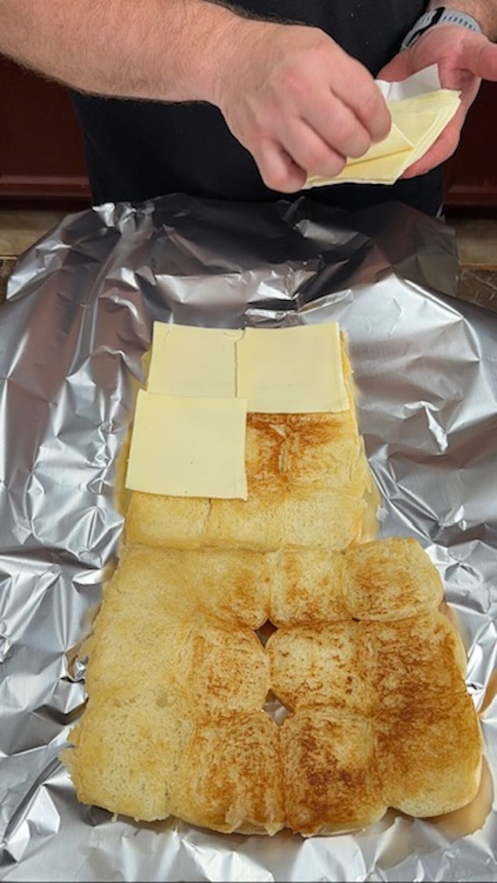A hand placing down slices of cheese on top of of toasted slider buns that are cut in half on top of foil.