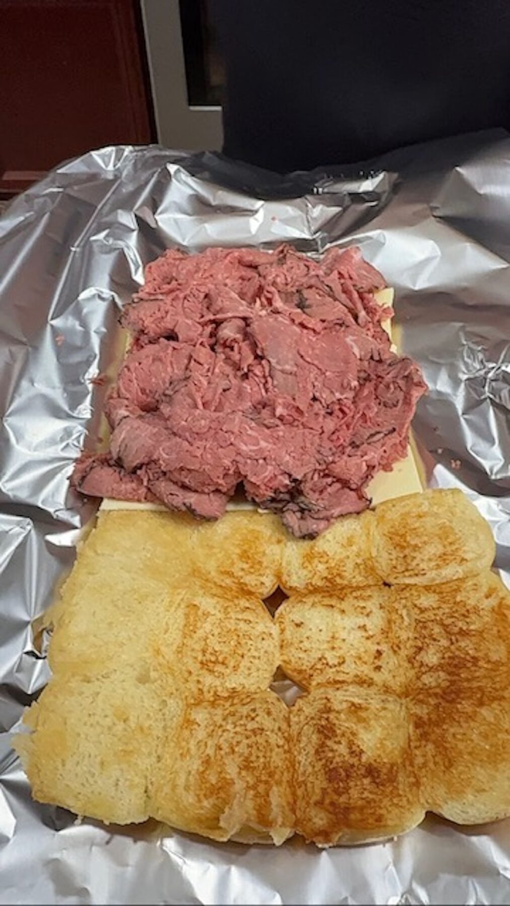 Shaved beef is placed on top of slider rolls layered with cheese on foil.