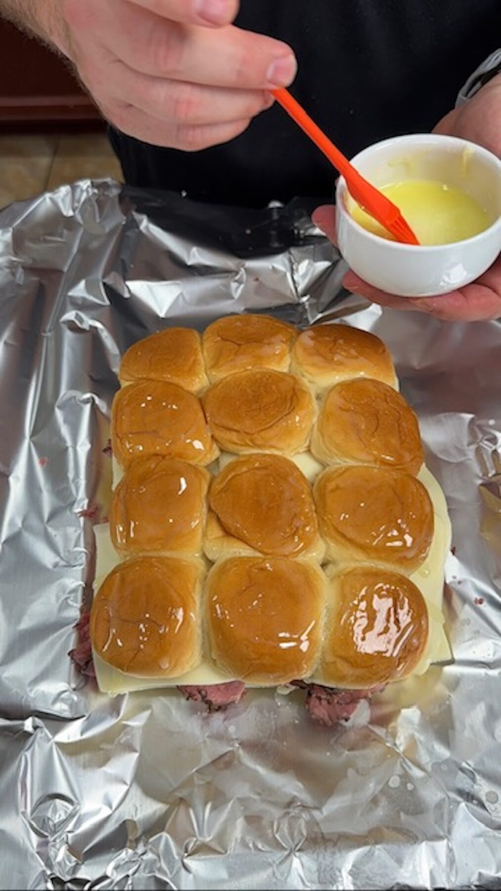 A hand is brushing melted butter on top of slider rolls on a piece of foil.
