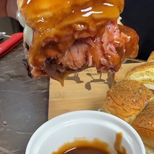 A hand is dipping a beef and cheese slider into a white ramekin of gravy.