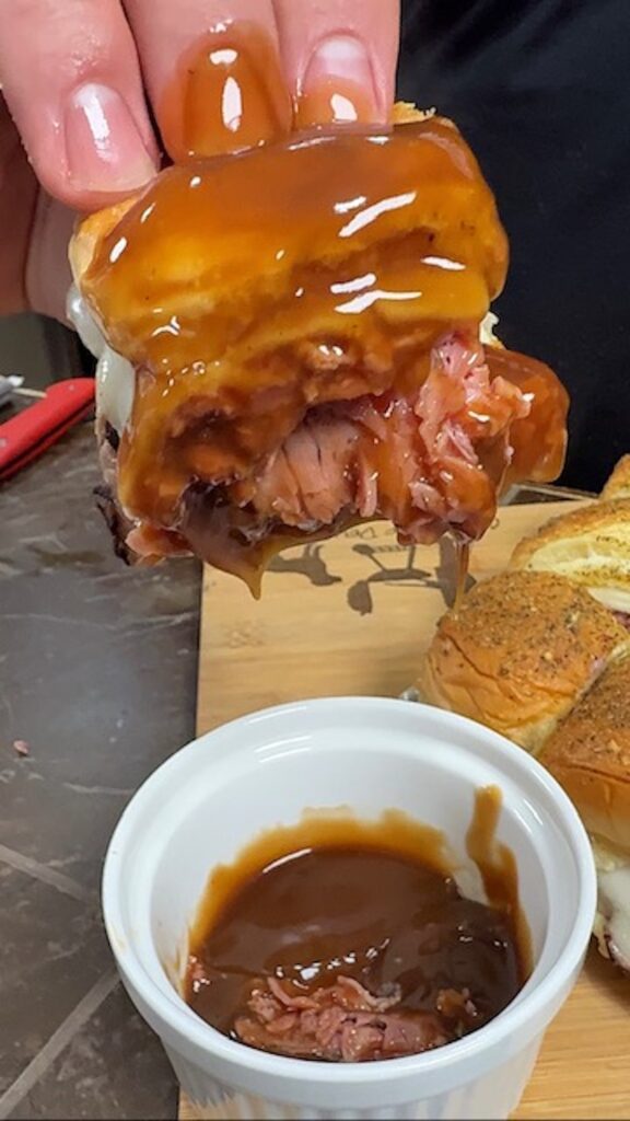 A hand is dipping a beef and cheese slider into a white ramekin of gravy.
