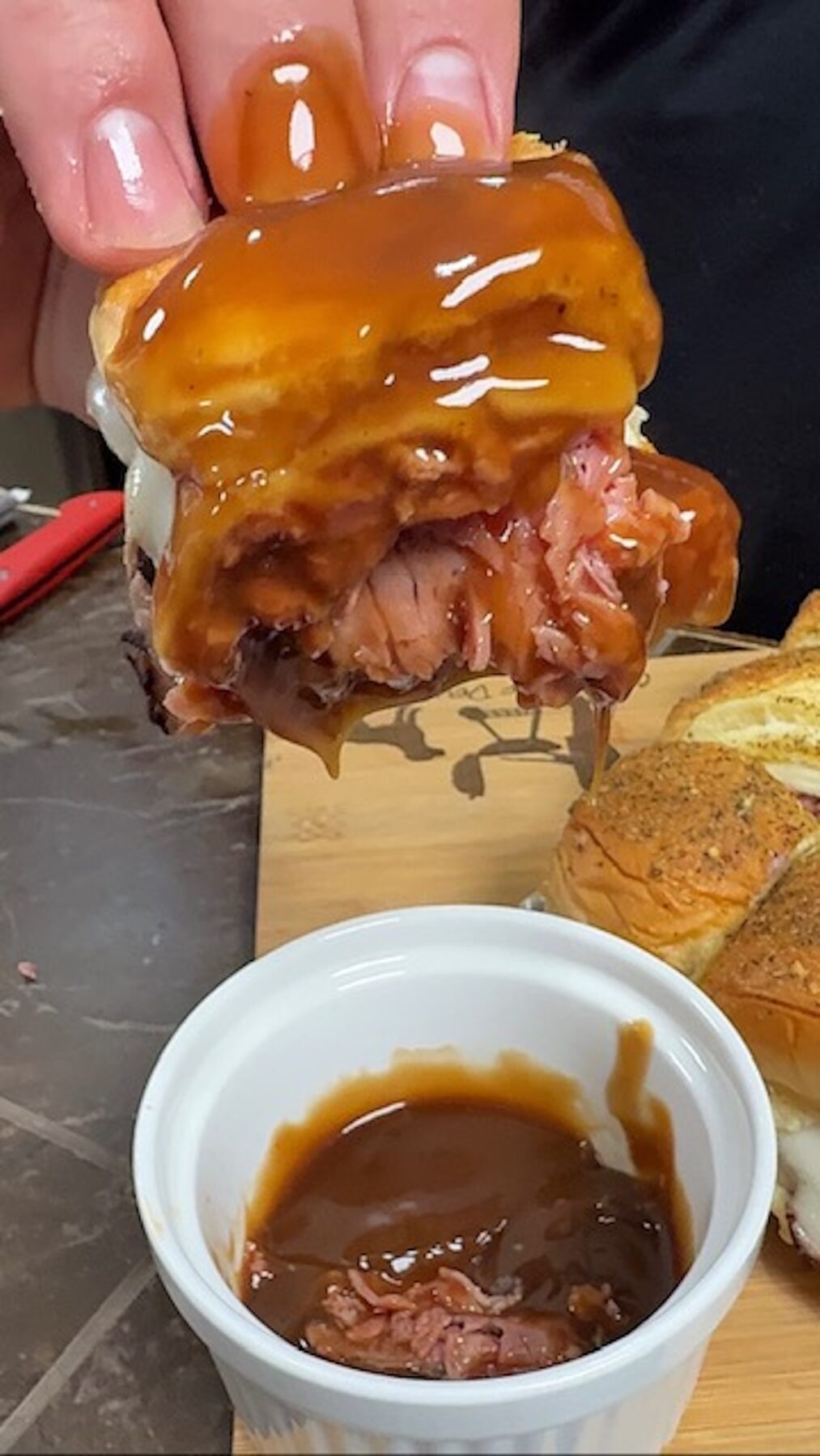 A hand is dipping a beef and cheese slider into a white ramekin of gravy.