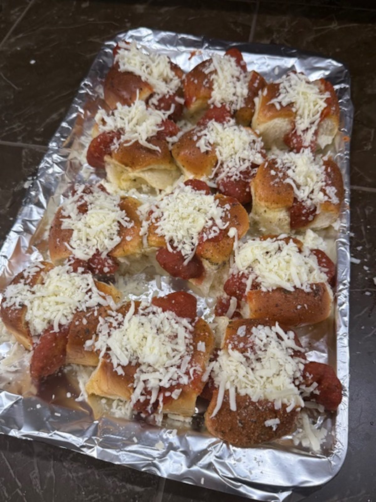 Shredded mozzarella is added on top of the chicken parm sliders on the foil-lined baking sheet.