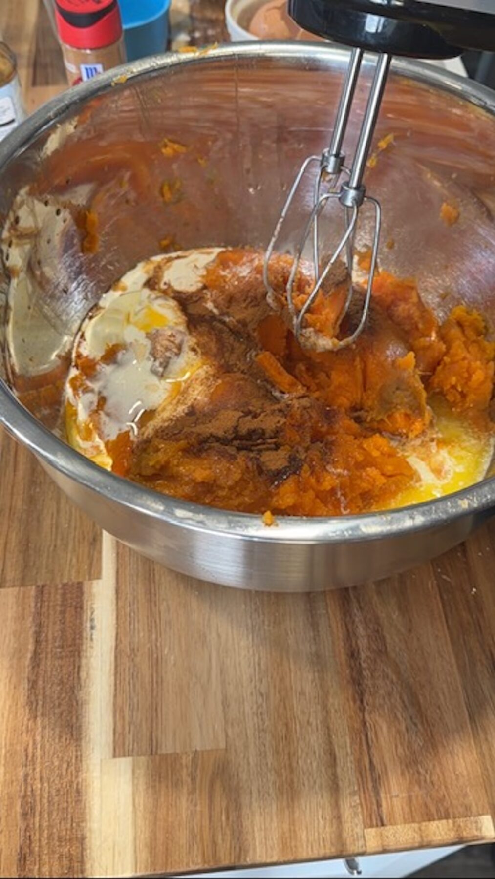 A metal bowl on a wooden cutting board with sweet potato casserole fillings and a hand mixer above.