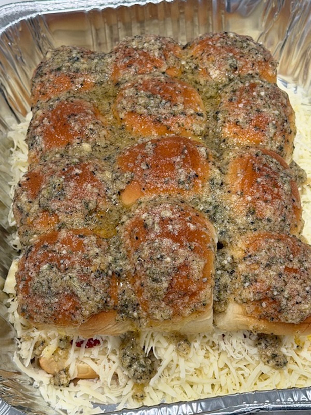 Top buns placed on top of pepperoni pizza sliders ingredients and brushed with garlic butter. The sliders are in a foil tray.