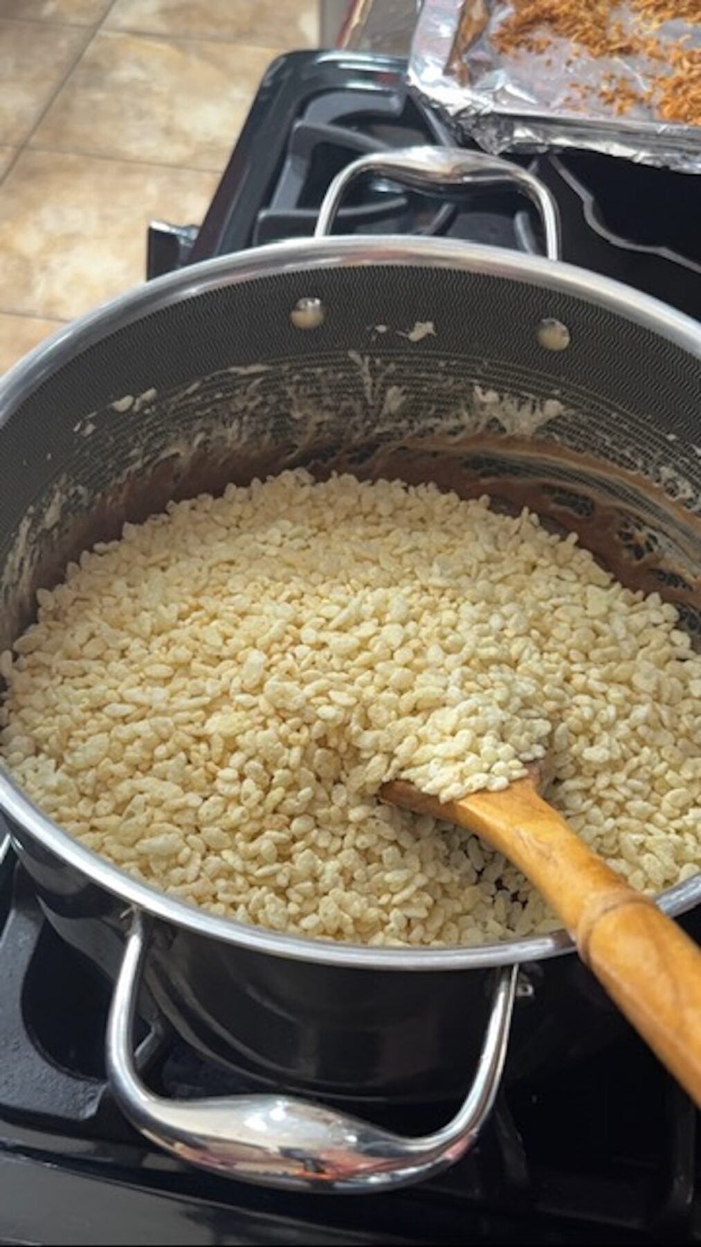A large pot with rice krispies, melted marshmallows, chocolate chips, and toasted coconut. A wooden spoon is resting on the side.