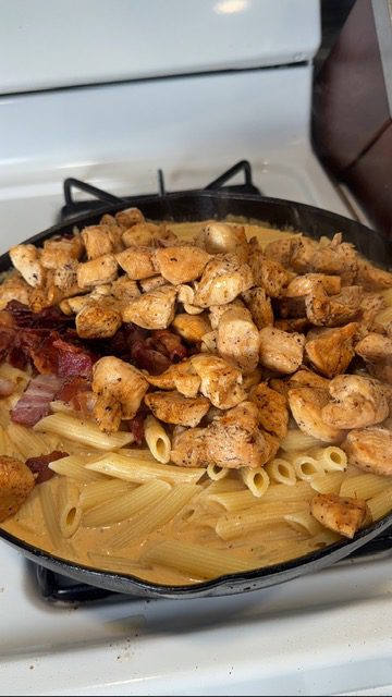 A cast iron skillet with crack chicken pasta, cooked chicken, bacon, and noodles on the stovetop.
