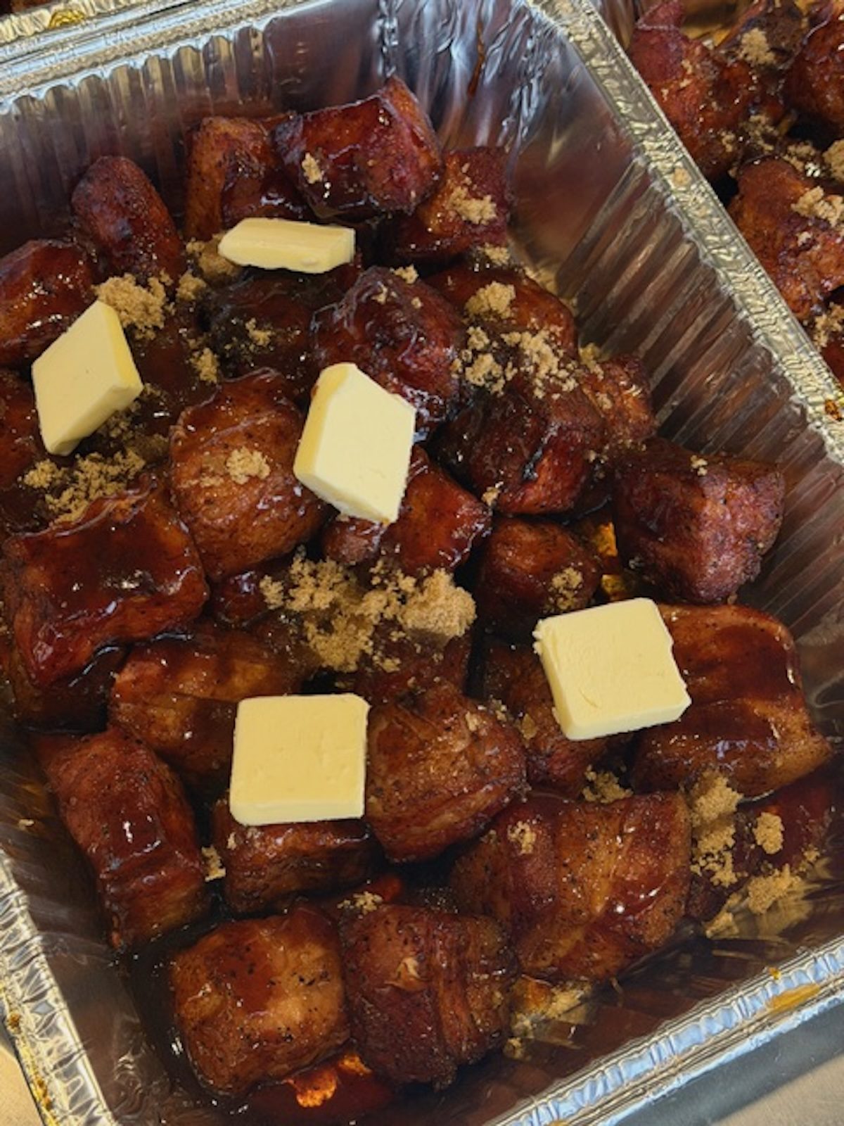Pork belly burnt ends in a foil tray with sugar and pats of butter on top.