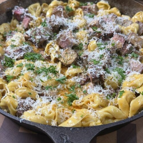 A cast iron with melted butter, tortellini, steak pieces, and shredded cheese.