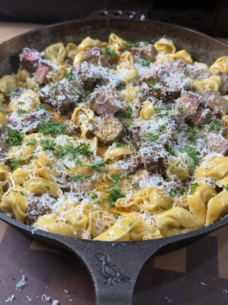 A cast iron with melted butter, tortellini, steak pieces, and shredded cheese.