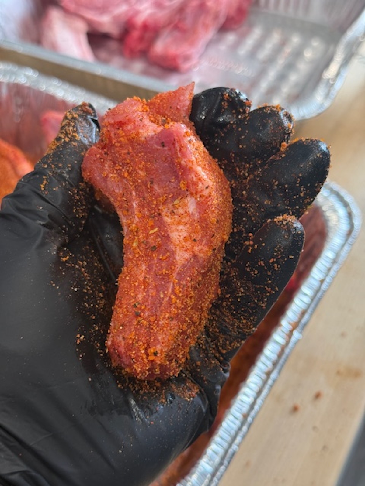 individual rib seasoned with bbq rub in hand wearing black glove. raw ribs in background in foil tray.