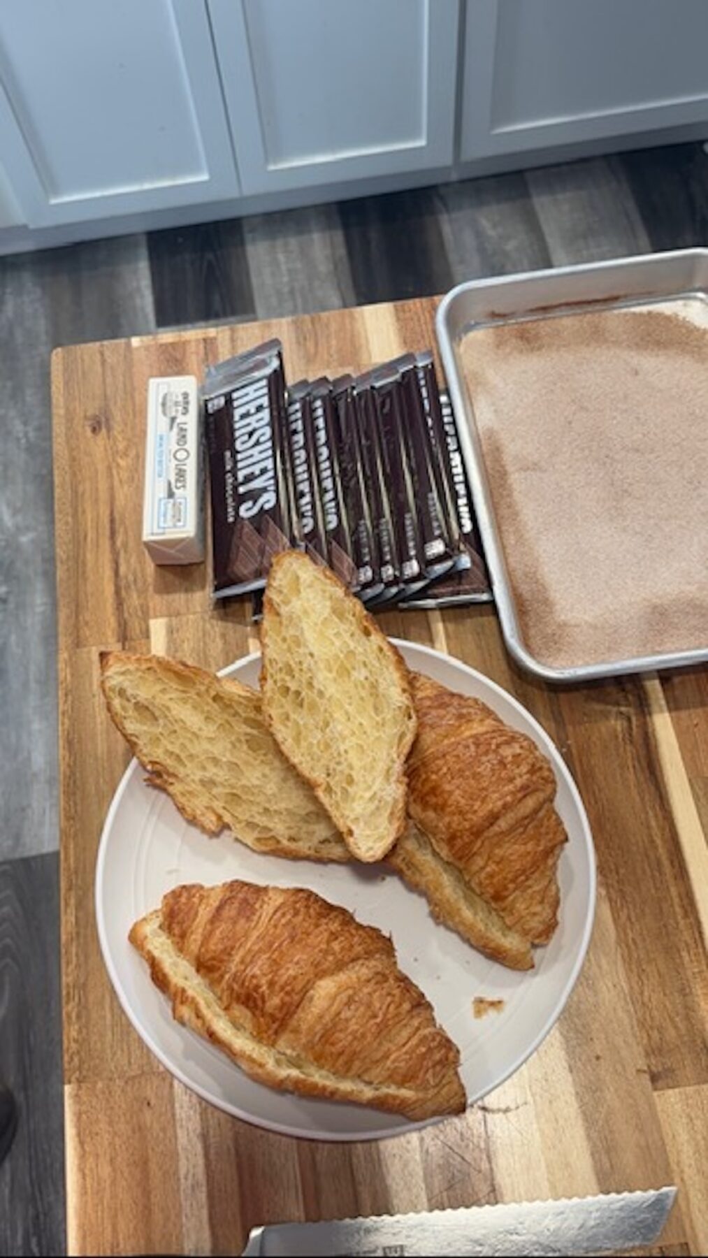 croissants halved on white plate next to chocolate bars and sheet pan with cinnamon sugar on it, all on cutting board
