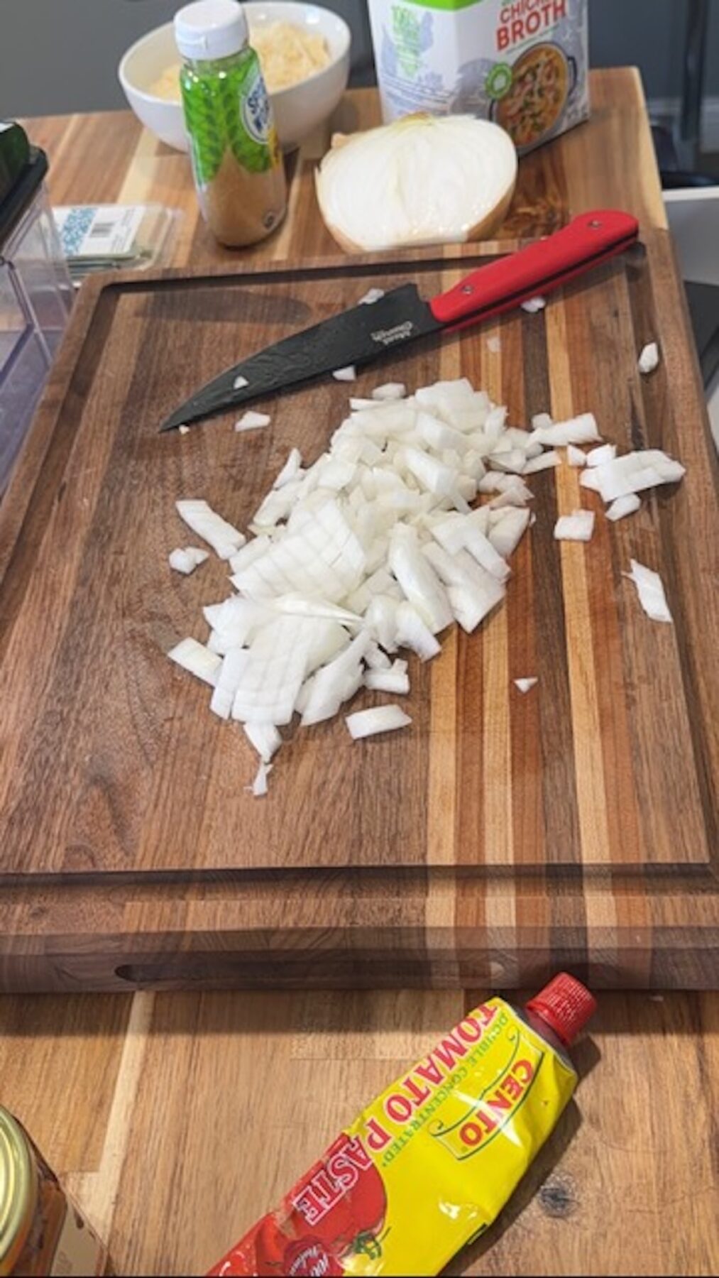 chipped onions next to tomato paste and knife on wooden cutting board. halved onion, broth and spices in background.
