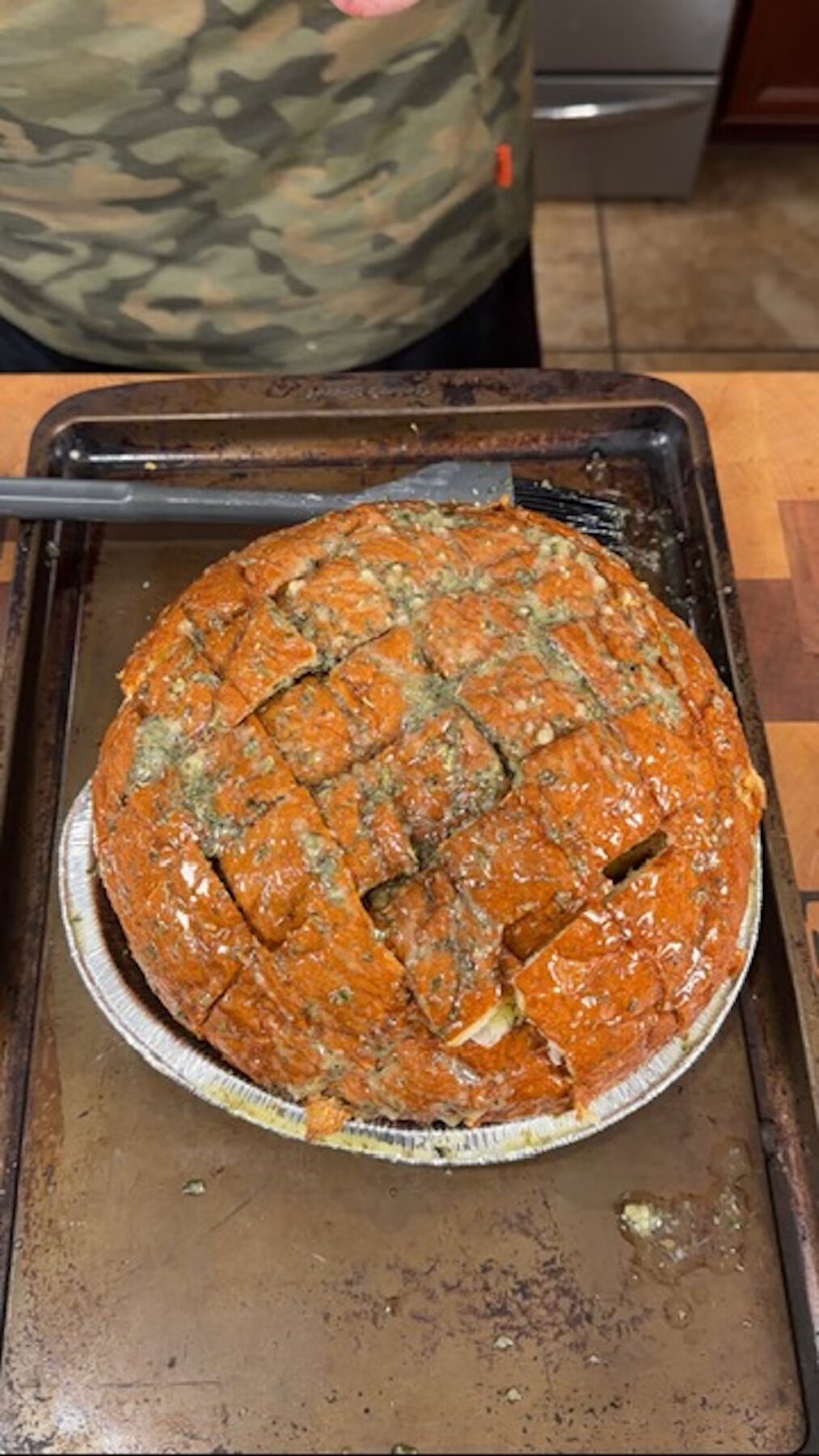 A loaf of bread with criss-cross cuts and melted herb butter all over it in a foil tin on a baking sheet.